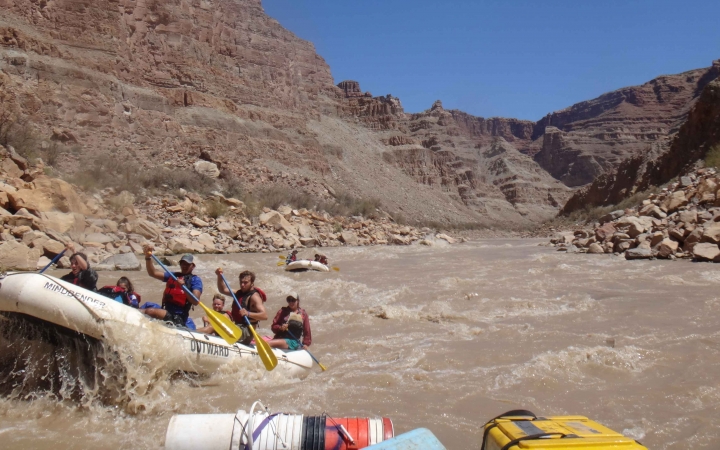 A group of students paddle a raft through whitewater. There is another raft in the background, and the river is framed by high canyon walls.